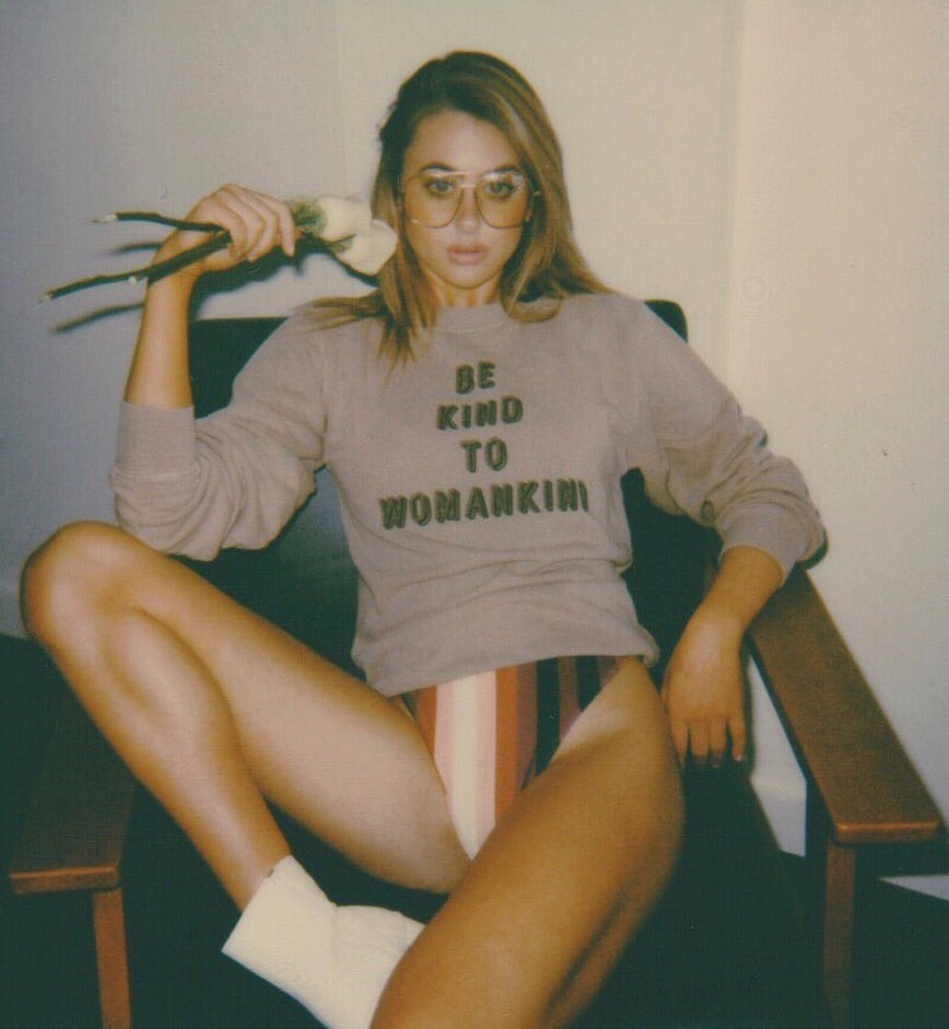 Woman sitting in chair wearing sandstone colored sweatshirt that says Be Kind to Womankind.