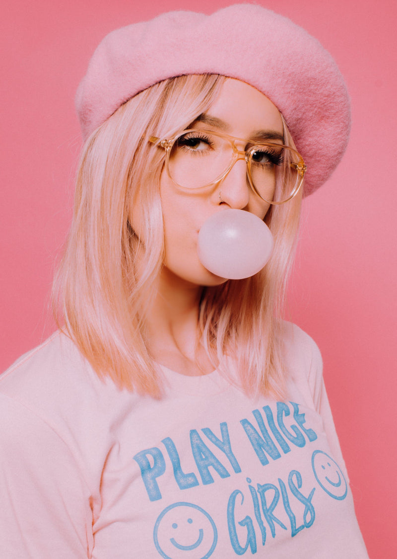 Girl blowing a bubble wearing Play Nice Girls soft pink cotton t-shirt by Top Knot Goods.