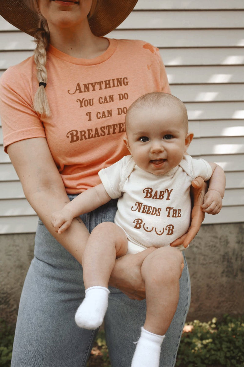 Mother holding baby that is wearing a funny baby onesie that says Baby Needs the Boob